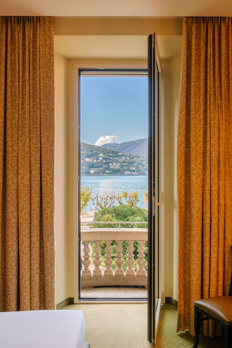 A hotel room with a large sliding glass door leading to a balcony with a view of a lake and mountains. - Palace Hotel (Como)